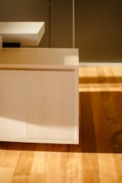 A modern wooden cabinet with straight lines and a light wood finish is partially illuminated by sunlight coming through a window. The light creates distinct shadows across the cabinet and the hardwood floor beneath it.