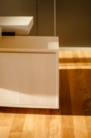 A modern wooden cabinet with straight lines and a light wood finish is partially illuminated by sunlight coming through a window. The light creates distinct shadows across the cabinet and the hardwood floor beneath it.