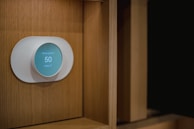 A modern, circular smart thermostat is mounted on a wooden wall. The device displays a message suggesting setup with Wi-Fi and shows the number 50, indicating an indoor temperature setting. The design appears sleek and minimalist.