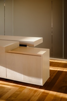 A modern, minimalist wooden cabinet with clean, sharp lines and a light wood finish. The cabinet is placed on a hardwood floor, with soft, natural light casting gentle shadows, enhancing the warm tones of the wood.