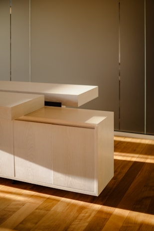 A modern, minimalist wooden cabinet with clean, sharp lines and a light wood finish. The cabinet is placed on a hardwood floor, with soft, natural light casting gentle shadows, enhancing the warm tones of the wood.