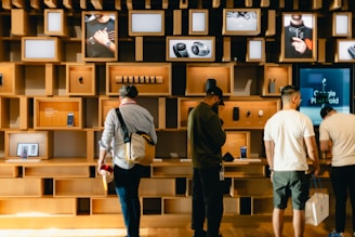 A group of people stand in front of a wall with various shelves displaying electronic devices and promotional images. The shelves are made of light wood, adding a modern and organized appearance. Some devices on display include smartphones and watches, with advertising screens showing relevant product images and information.