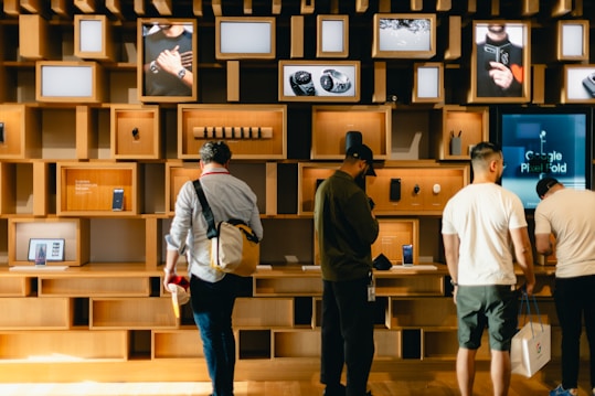 A group of people stand in front of a wall with various shelves displaying electronic devices and promotional images. The shelves are made of light wood, adding a modern and organized appearance. Some devices on display include smartphones and watches, with advertising screens showing relevant product images and information.
