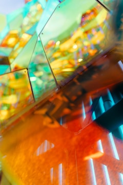 A vibrant and abstract composition featuring reflections and refractions through colorful glass or acrylic panels. The overlapping panels create a kaleidoscope effect with hues of green, blue, and orange. The image has a modern and artistic aesthetic with a play of light and shadows.