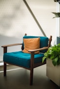 A modern wooden armchair with a teal cushion is placed near a large plant in a pot. An orange pillow is centered on the chair, and the setting includes a light-colored wall with subtle patterns. Natural light softly illuminates the scene.