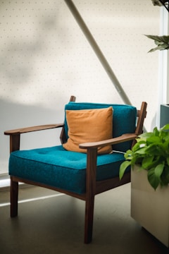 A modern wooden armchair with a teal cushion is placed near a large plant in a pot. An orange pillow is centered on the chair, and the setting includes a light-colored wall with subtle patterns. Natural light softly illuminates the scene.