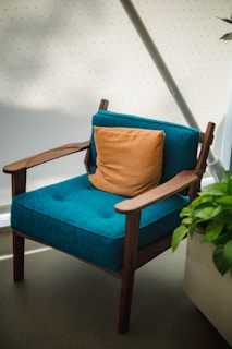 A modern wooden armchair features teal upholstery and an orange cushion. The setup includes a lush green plant beside the chair, complemented by a minimalist background with a soft dot pattern. Ambient lighting gives a cozy and inviting feel.