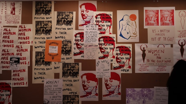 A collage of protest posters is displayed on the wall, featuring bold texts advocating for women's rights. The dominant messages include phrases like 'Women's Liberation' and 'Women Need Not Always Keep Their Mouths Shut.' There are artistic renderings of human figures and parts of slogans that focus on women's empowerment and anti-war sentiments.