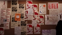 A collage of protest posters is displayed on the wall, featuring bold texts advocating for women's rights. The dominant messages include phrases like 'Women's Liberation' and 'Women Need Not Always Keep Their Mouths Shut.' There are artistic renderings of human figures and parts of slogans that focus on women's empowerment and anti-war sentiments.