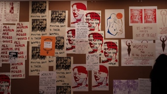 A collage of protest posters is displayed on the wall, featuring bold texts advocating for women's rights. The dominant messages include phrases like 'Women's Liberation' and 'Women Need Not Always Keep Their Mouths Shut.' There are artistic renderings of human figures and parts of slogans that focus on women's empowerment and anti-war sentiments.