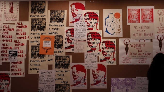 A collage of protest posters is displayed on the wall, featuring bold texts advocating for women's rights. The dominant messages include phrases like 'Women's Liberation' and 'Women Need Not Always Keep Their Mouths Shut.' There are artistic renderings of human figures and parts of slogans that focus on women's empowerment and anti-war sentiments.