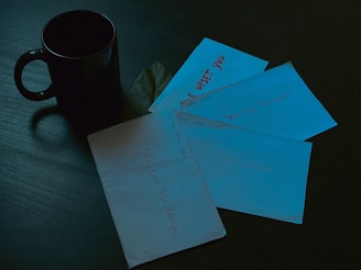 A dimly lit scene features a black coffee mug on a dark wooden table. Surrounding the mug are multiple envelopes, each inscribed with personal messages such as 'We don't talk anymore', 'You feel alone', 'Open when you're upset', and 'You can't fail forever'. The envelopes and writing create a somber atmosphere.