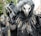 A person is dressed in a dark, elaborate costume resembling a bird or mythical creature, featuring a long beak-like mask, hood, and black feathers. The costume includes twig-like decorations around the shoulders and black gloves wrapped around a wooden staff.