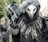 A person is dressed in a dark, elaborate costume resembling a bird or mythical creature, featuring a long beak-like mask, hood, and black feathers. The costume includes twig-like decorations around the shoulders and black gloves wrapped around a wooden staff.