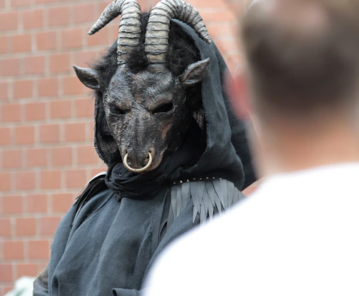 Obsidian black background with faint blood-red veins, framing a silhouette of the goat-like mask in mist.