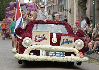 A vibrant parade float in the shape of a classic car, adorned with red and white flowers and featuring the words 'Cuba' and 'Libre.' Several children, some holding trophies, are standing on the float, which is moving down a street lined with onlookers. A large flag is displayed alongside the car.