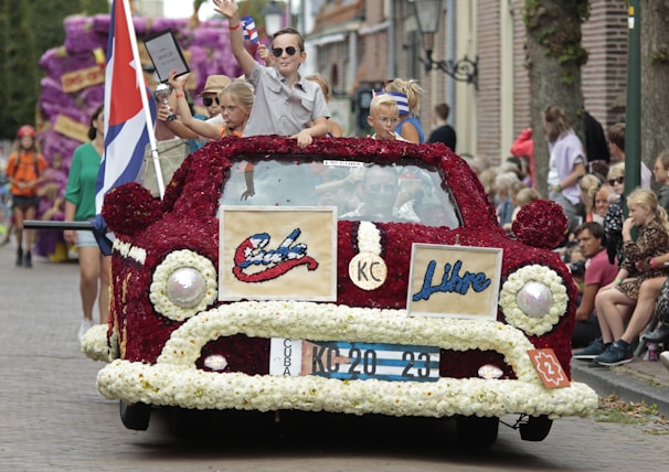A vibrant parade float in the shape of a classic car, adorned with red and white flowers and featuring the words 'Cuba' and 'Libre.' Several children, some holding trophies, are standing on the float, which is moving down a street lined with onlookers. A large flag is displayed alongside the car.