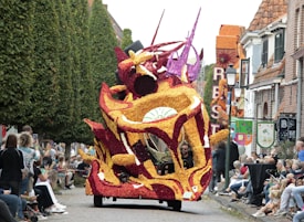 A vibrant, complex float covered in colorful flowers drives down a street lined with spectators. The float features swirling patterns and abstract designs, primarily in shades of red, yellow, and white. Tall trees and brick buildings flank the parade route.