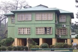 A three-story building with a green exterior and multiple windows. The structure is surrounded by well-maintained bushes and trees. Several air conditioning units are mounted on the building walls.