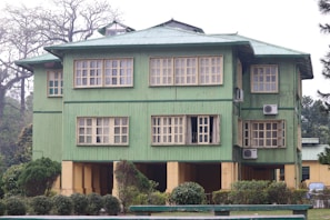 A three-story building with a green exterior and multiple windows. The structure is surrounded by well-maintained bushes and trees. Several air conditioning units are mounted on the building walls.