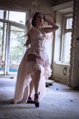 A woman dressed in a flowing, vintage-style gown stands gracefully inside an abandoned, dimly lit room. The decor is rustic and distressed, with scattered debris on the floor, adding a haunting, ethereal atmosphere. She wears bright purple ballet shoes and a matching headpiece, and her pose is elegant, as if striking a ballet pose.