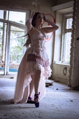 A woman dressed in a flowing, vintage-style gown stands gracefully inside an abandoned, dimly lit room. The decor is rustic and distressed, with scattered debris on the floor, adding a haunting, ethereal atmosphere. She wears bright purple ballet shoes and a matching headpiece, and her pose is elegant, as if striking a ballet pose.