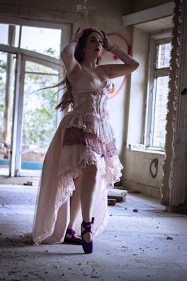 A woman dressed in a flowing, vintage-style gown stands gracefully inside an abandoned, dimly lit room. The decor is rustic and distressed, with scattered debris on the floor, adding a haunting, ethereal atmosphere. She wears bright purple ballet shoes and a matching headpiece, and her pose is elegant, as if striking a ballet pose.