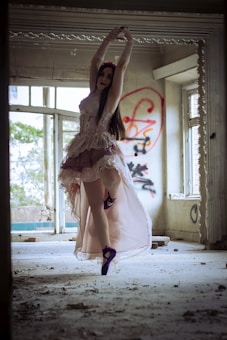 A ballet dancer in a flowing, light pink dress performs a graceful pose in an abandoned, graffiti-covered room. Dust and debris are scattered on the floor, and natural light streams through large windows, casting soft illumination on the scene.