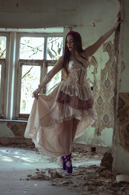 A woman in an elegant, flowing dress stands in a dilapidated, abandoned room with broken plaster and peeling wallpaper. Natural light filters through large, dirty windows, casting soft shadows on the floor. Her dress features intricate lace details and she is wearing bright purple ballet shoes. Her pose is graceful and poised, contrasting with the rough surroundings.