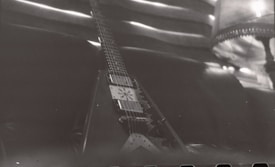 A monochrome image of a V-shaped electric guitar resting against a dark leather couch. The guitar features visible pickups and strings, with its neck extending into the background. In the upper right corner, there's a part of a lamp with a fringed lampshade and some soft lighting.