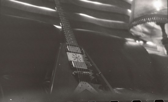 A monochrome image of a V-shaped electric guitar resting against a dark leather couch. The guitar features visible pickups and strings, with its neck extending into the background. In the upper right corner, there's a part of a lamp with a fringed lampshade and some soft lighting.