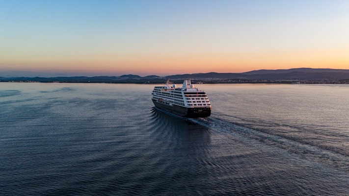 A large cruise ship is sailing on a calm sea during a picturesque sunset. The vessel is moving toward the distant coastline, which is dotted with gentle hills. The sky displays a gradient of colors, transitioning from pale blue to warm orange hues.