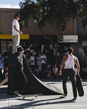 A group of skateboarders gathers around a small ramp in an urban setting. One person stands atop the ramp, while another is positioned at its base on a skateboard. A third individual walks nearby, carrying a skateboard. Spectators sit and stand around, watching the activity. The scene takes place under a tree with ample sunlight creating shadows.