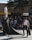 A group of skateboarders gathers around a small ramp in an urban setting. One person stands atop the ramp, while another is positioned at its base on a skateboard. A third individual walks nearby, carrying a skateboard. Spectators sit and stand around, watching the activity. The scene takes place under a tree with ample sunlight creating shadows.