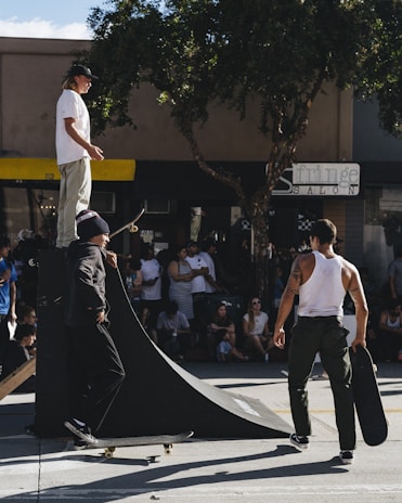 A group of skateboarders gathers around a small ramp in an urban setting. One person stands atop the ramp, while another is positioned at its base on a skateboard. A third individual walks nearby, carrying a skateboard. Spectators sit and stand around, watching the activity. The scene takes place under a tree with ample sunlight creating shadows.