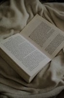 An open book with pages gently turning, resting on a beige linen cloth with olive accents.