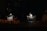 A small street food stall is illuminated by a solitary light, showcasing a vendor working at night. There are several stools arranged in front of the stall, which has signs in Vietnamese advertising its offerings. The scene captures a quiet, nighttime atmosphere.