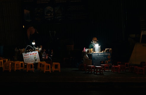 A small street food stall is illuminated by a solitary light, showcasing a vendor working at night. There are several stools arranged in front of the stall, which has signs in Vietnamese advertising its offerings. The scene captures a quiet, nighttime atmosphere.