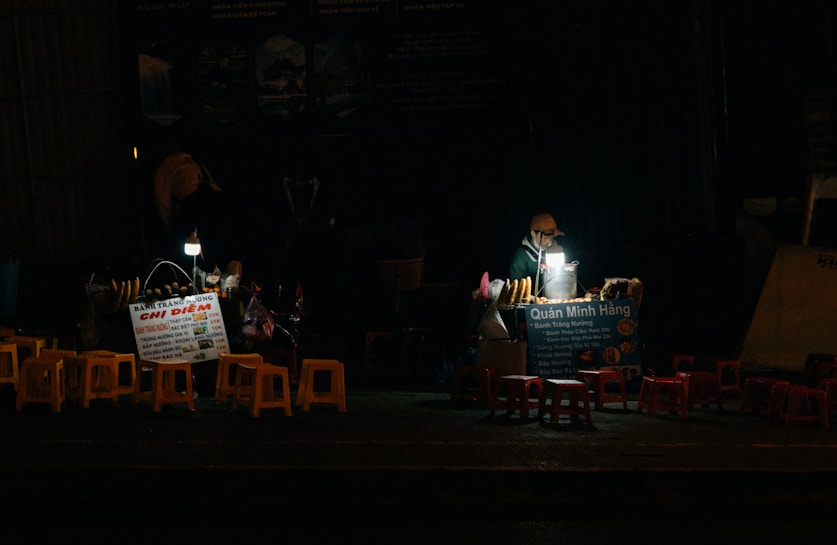 A small street food stall is illuminated by a solitary light, showcasing a vendor working at night. There are several stools arranged in front of the stall, which has signs in Vietnamese advertising its offerings. The scene captures a quiet, nighttime atmosphere.