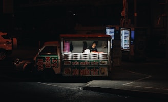 A nighttime scene featuring a small street food vendor stall with a person inside preparing or selling food. The stall is lit from within, drawing focus to the various pots and containers with what appears to be Vietnamese lettering on the signage. The surrounding area is dimly lit, with vending machines visible in the background, and a parked car partially seen to the left.