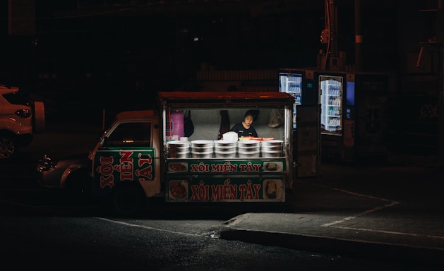 A nighttime scene featuring a small street food vendor stall with a person inside preparing or selling food. The stall is lit from within, drawing focus to the various pots and containers with what appears to be Vietnamese lettering on the signage. The surrounding area is dimly lit, with vending machines visible in the background, and a parked car partially seen to the left.