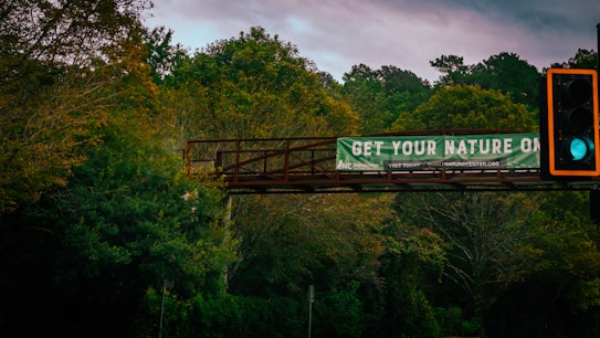 A lush and densely wooded area with a metal pedestrian bridge crossing over. The bridge supports a banner that reads 'Get Your Nature On' and promotes a nature center website. In the foreground, a traffic light is visible with a green light illuminated against the backdrop of trees.