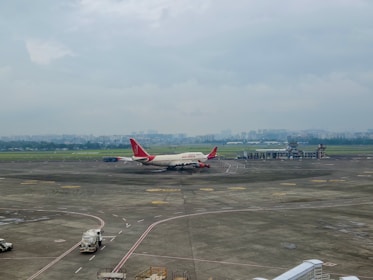 A commercial airplane with red and white livery sits on the tarmac at an airport. The background includes hangars and a city skyline under a cloudy sky. Ground service vehicles are visible near the plane, and runways are marked clearly on the pavement.