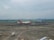 A commercial airplane with red and white livery sits on the tarmac at an airport. The background includes hangars and a city skyline under a cloudy sky. Ground service vehicles are visible near the plane, and runways are marked clearly on the pavement.