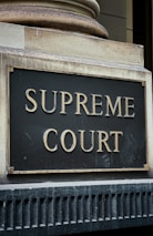 A close-up view of a sign with the words 'Supreme Court' in gold-colored letters mounted on a textured gray and black background. The sign is bordered by an aged metal frame and positioned on an architectural feature with classical detailing.