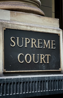 A close-up view of a sign with the words 'Supreme Court' in gold-colored letters mounted on a textured gray and black background. The sign is bordered by an aged metal frame and positioned on an architectural feature with classical detailing.