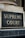 A close-up view of a sign with the words 'Supreme Court' in gold-colored letters mounted on a textured gray and black background. The sign is bordered by an aged metal frame and positioned on an architectural feature with classical detailing.