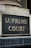 A close-up view of a sign with the words 'Supreme Court' in gold-colored letters mounted on a textured gray and black background. The sign is bordered by an aged metal frame and positioned on an architectural feature with classical detailing.