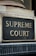 A close-up view of a sign with the words 'Supreme Court' in gold-colored letters mounted on a textured gray and black background. The sign is bordered by an aged metal frame and positioned on an architectural feature with classical detailing.