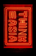 Bright red neon sign with stylized lettering, possibly spelling out 'Turn Up Asia'. The sign is enclosed in a rectangular frame against a dark background.